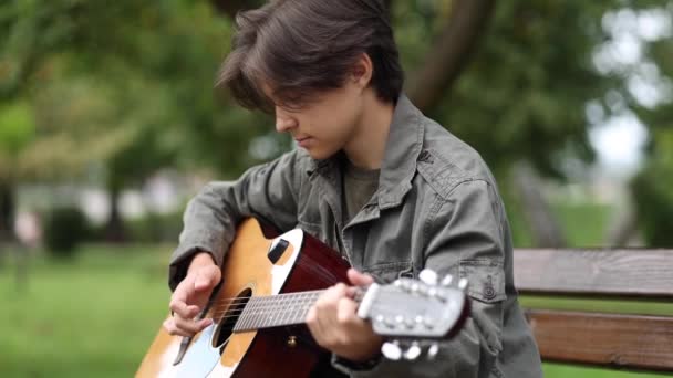 Teenage boy sedí na lavičce a hraje na kytaru venku v parku. Dlouhán sám hraje na kytaru. Podzimní nálada — Stock video