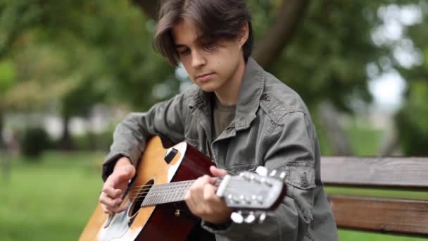 Teenage boy sedí na lavičce a hraje na kytaru venku v parku. Dlouhán sám hraje na kytaru. Podzimní nálada