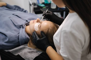 Güzellik stüdyosunda kirpik yapan bir kadın. Sarı saç modelinin kirpikleri için boyası, kıvırcığı, kaplaması ve uzantısı var. Siyah eldivenler