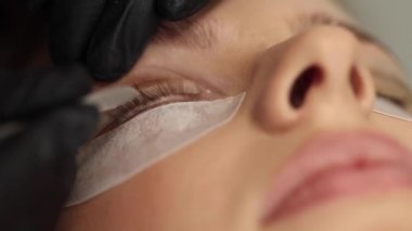 Kirpik tedavisi prosedürlerine yakın çekim. Güzel bir kadın, sarı saç modeli kirpikleri var. Kadın kirpikleri boyuyor, kıvırıyor, kaplıyor ve uzatıyor. Makro video