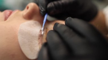 Kirpik tedavisi prosedürlerine yakın çekim. Güzel bir kadın, sarı saç modeli kirpikleri var. Kadın kirpikleri boyuyor, kıvırıyor, kaplıyor ve uzatıyor. Yan görünüm