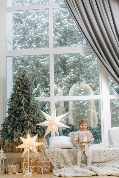 快乐美丽的女孩在大窗户前接受礼物。圣诞节的时候外面下雪了。圣诞树 — 图库照片