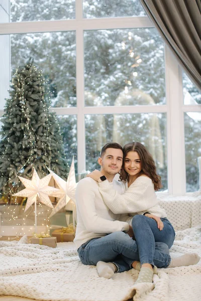 Un chico guapo con su hermosa novia se abrazan en casa en la víspera de Navidad. Ajuste árbol y nieve outsif. Humor de Navidad — Foto de Stock