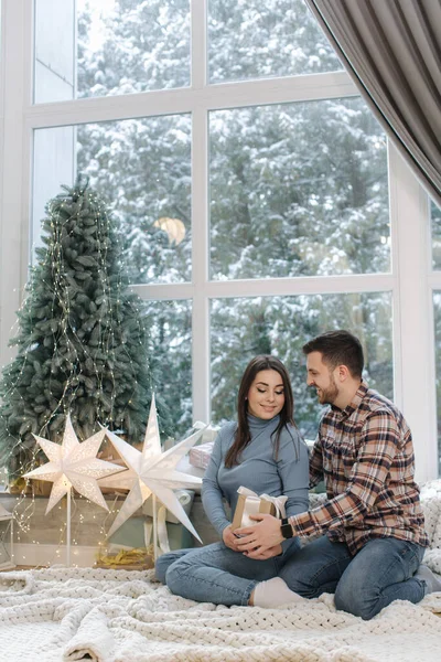 Jovem atraente com seu amado marido segurar presente de Natal. Casal sentado na frente de uma grande janela e perto do abeto. Neve lá fora — Fotografia de Stock