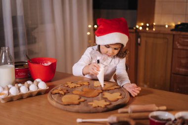 Noel Baba şapkalı küçük kız evde zencefilli ekmek süslüyor. Noel ve Yeni Yıl gelenekleri. Noel pastanesi. Mutlu tatiller.
