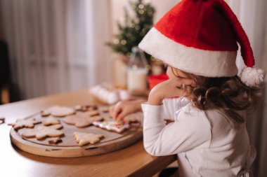 Noel Baba şapkalı küçük kız evde zencefilli ekmek süslüyor. Noel ve Yeni Yıl gelenekleri. Noel pastanesi. Mutlu tatiller.