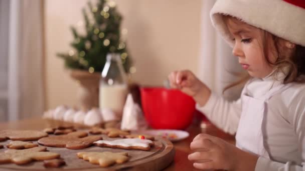Niña en sombrero de santa adorna pan de jengibre en casa. Concepto de tradiciones de Navidad y Año Nuevo. Humor de Navidad — Vídeos de Stock