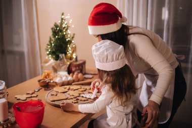 Noel Baba şapkalı anne küçük kızıyla zencefilli ekmeği jöleli süsledi. Noel ve Yeni Yıl gelenekleri. Noel pastanesi. İyi tatiller.