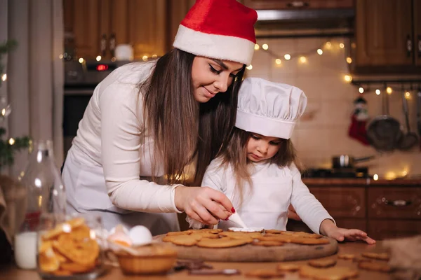 Noel Baba şapkalı anne küçük kızıyla zencefilli ekmeği jöleli süsledi. Noel ve Yeni Yıl gelenekleri. Noel pastanesi. İyi tatiller.