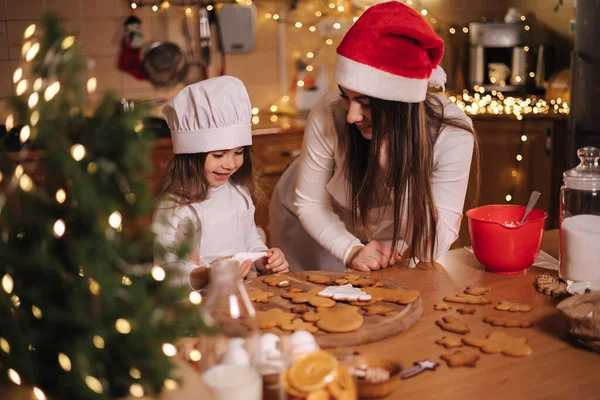 Güzel annesiyle mutlu küçük kız evde zencefilli ekmek yapıyor. Mutfakta Noel süsü. Peri ışıklarıyla fidan ağacı
