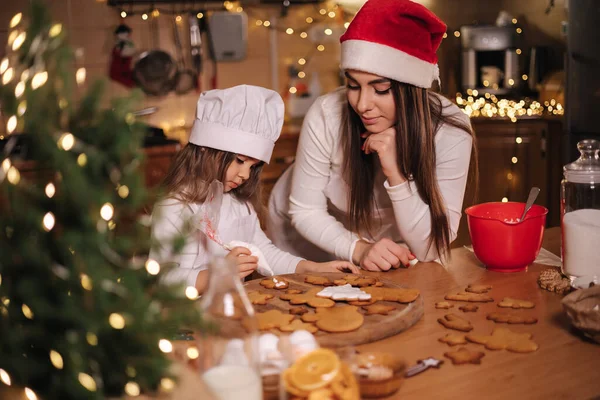 Güzel annesiyle mutlu küçük kız evde zencefilli ekmek yapıyor. Mutfakta Noel süsü. Peri ışıklarıyla fidan ağacı
