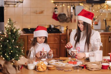 Kadın ve küçük kızı mutfakta lokumla lezzetli kakao içiyorlar. Noel arifesinde ev dekorasyonu. Annem ve Noel Baba şapkalı küçük kız. Peri ışıklarının arka planı. Ağaca sığdır