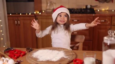 Kırmızı Noel Baba şapkalı sevimli küçük kız masanın yanında dans ediyor. Evde Noel için hazırlanıyorum. Zencefilli çörek.