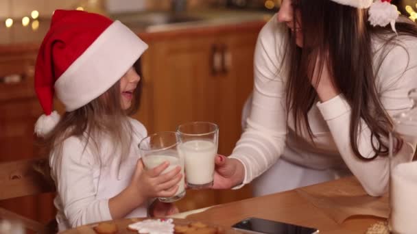 Hermosa niña con su madre en sombrero de santa sentado junto a la mesa en frente de la cocina decorada luces de hadas y beber leche fresca. Humor de Navidad — Vídeo de stock