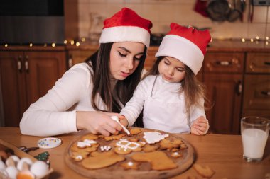Noel Baba şapkalı anne küçük kızıyla zencefilli ekmeği jöleli süsledi. Noel ve Yeni Yıl gelenekleri. Noel pastanesi. İyi tatiller.