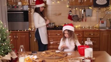 Güzel annesiyle mutlu küçük kız evde zencefilli ekmek yapıyor. Mutfakta Noel süsü. Peri ışıklarıyla fidan ağacı