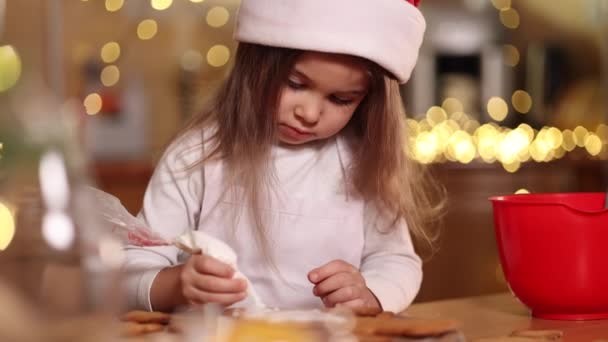 Menina em chapéu de santas decora pão de gengibre usando esmalte branco. Conceito de tradições de Natal e Ano Novo. Padaria de Natal. Boas festas — Vídeo de Stock