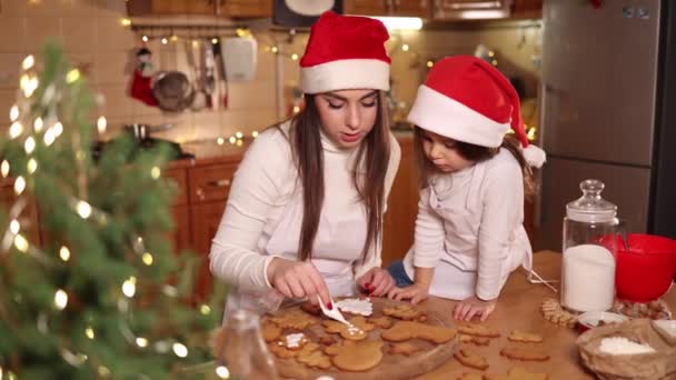 Menina feliz com sua bela mãe fazer pão de gengibre em casa. Decoração de Natal na cozinha. Abeto com luzes de fadas — Vídeo de Stock