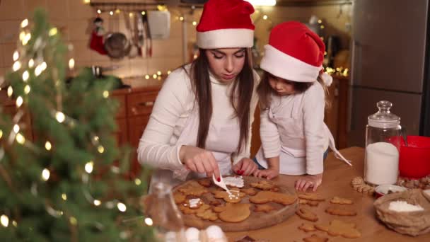 Putri kecil yang manis dengan ibunya di dapur selama liburan Natal. Pohon yang cocok dengan dekorasi di meja. Gadis membuat roti jahe. Gadis bertopi santa — Stok Video