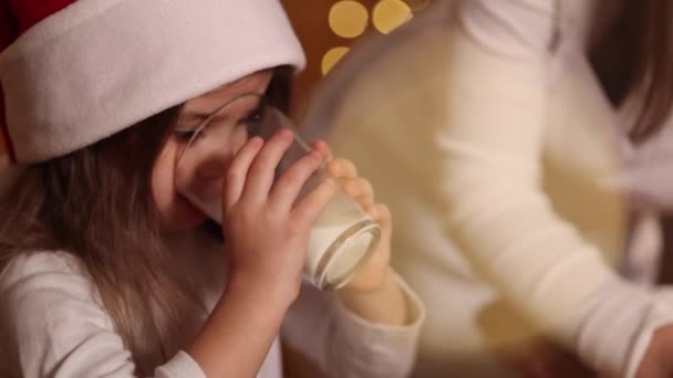 Krásná holčička se svou matkou v Santa klobouku sedí u stolu před kuchyní zdobené víla světla a pít čerstvé mléko. vánoční nálada — Stock video
