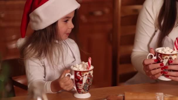 Nainen ja hänen tyttärensä juovat kaakaota vaahtokarkin kanssa keittiössä. Koti sisustettu jouluaattona. Äiti ja pikkutyttö jouluhatussa. Taustaa keijuvalot. Sovita puu. lähikuva — kuvapankkivideo