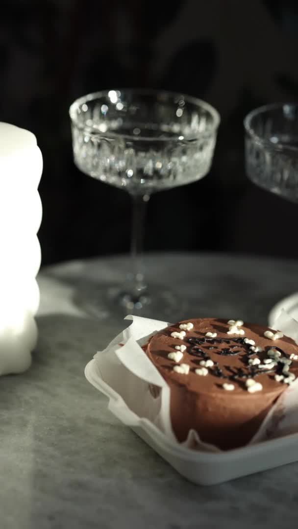 Шоколадный пирог в экологической коробке с деревянной ложкой. Белая свеча и бокалы для шампанского на роскошном мраморном столе. Вертикальное видео 4K 50fps — стоковое видео