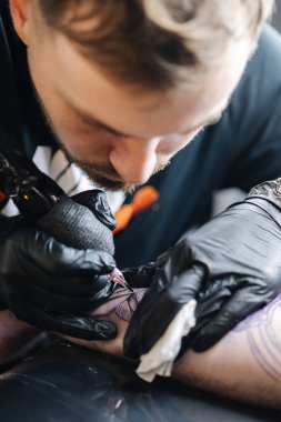 Genç bir dövmeci, kablosuz dövme makinesinin iğnesini kullanarak cildine siyah mürekkep sokuyor. Elinde dövme var..