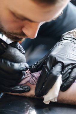 Genç bir dövmeci, kablosuz dövme makinesinin iğnesini kullanarak cildine siyah mürekkep sokuyor. Elinde dövme var..