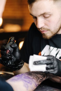 Genç bir dövmeci, dövme makinesinin iğnesini kullanarak cildine siyah mürekkep bulaştırıyor. Elinde dövme var.