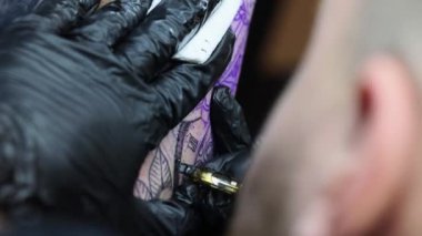 Genç bir dövmeci, kablosuz dövme makinesinin iğnesini kullanarak cildine siyah mürekkep sokuyor. Elinde dövme var. Dikey video