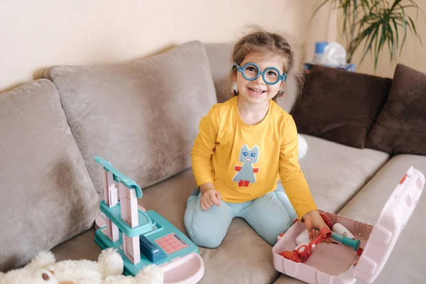 Imádnivaló kislány mosolyog és doktorosdit játszik a szobájában. Aranyos hároméves lány játszadozik. A lányok sárga blúzt és kék nadrágot viselnek. Ukrán aranyos gyerek — Stock Fotó