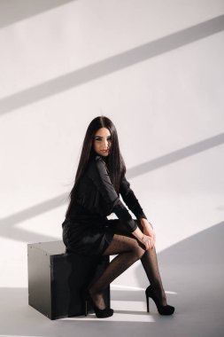 Siyah elbiseli zarif bir kadın siyah meydanda oturuyor. Stüdyo fotoğraf çekiminde esmer bir kadın. Güneşten gelen ışık beyaz arkaplanda bir gölge oluşturur.