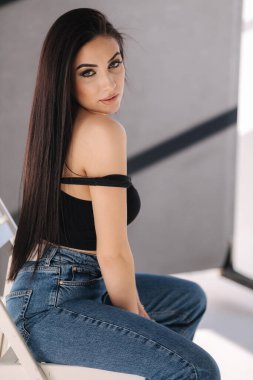Siyah başlıklı seksi kadın ve kot pantolon stüdyo fotoshoots sırasında sandalyede oturuyor. Esmer kız fotoğrafçıya poz veriyor. Beyaz arkaplanda gölge