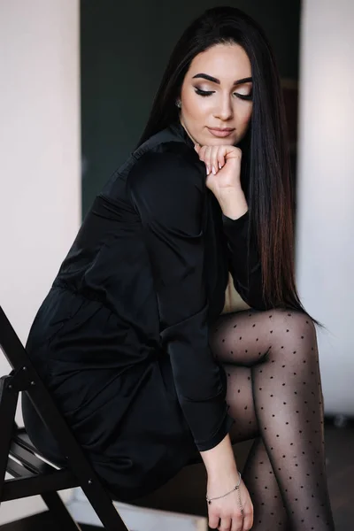 Frumoasă brunetă femeie în rochie neagră elegantă. Femeie așezată pe scaun negru — Fotografie, imagine de stoc
