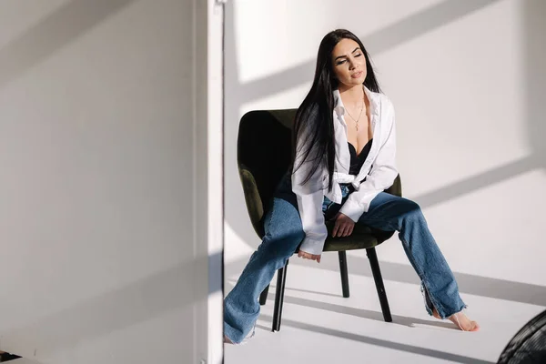 Pretty woman has photosession in studio. Sexy girl in denim and white shirt. Barefoot woman. Light from the sun makes a shadow on a white background. Stylish green chair — ストック写真