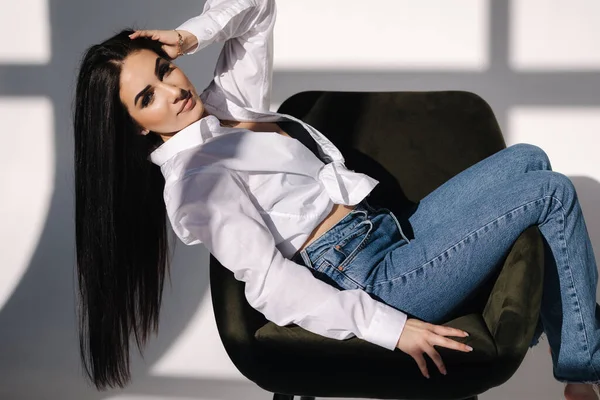Female sitting on green chair. Her black hair is hanging down. Pretty woman in denim and white shirt — стоковое фото