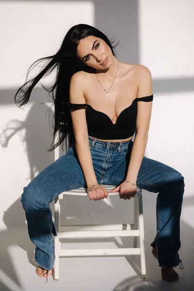 Mulher sexy em preto tópico e jeans azul senta-se na cadeira durante photoshoots estúdio. Menina morena posando para fotógrafo. Sombra sobre fundo branco — Fotografia de Stock