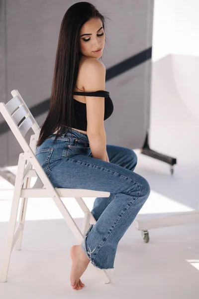 Mulher sexy em preto tópico e jeans azul senta-se na cadeira durante photoshoots estúdio. Menina morena posando para fotógrafo. Sombra sobre fundo branco — Fotografia de Stock