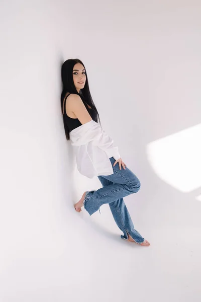 Pretty woman barefoot in studio. Beautiful brunette girl in black topic with white shirt and blue denim jeans. White background of cyclorama — ストック写真