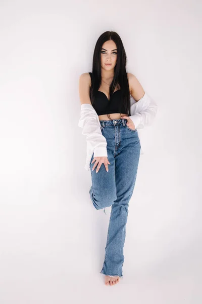 Pretty woman barefoot in studio. Beautiful brunette girl in black topic with white shirt and blue denim jeans. White background of cyclorama —  Fotos de Stock
