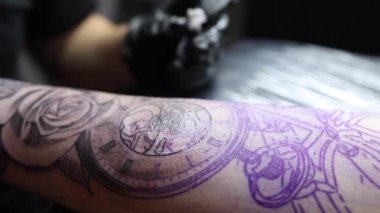 Genç bir dövmeci, kablosuz dövme makinesinin iğnesini kullanarak cildine siyah mürekkep sokuyor. Elinde dövme var.