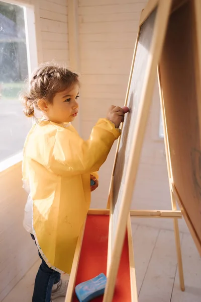 Young artist paints on wooden boards with chalk. Cute girl in yeallow raincoat — Foto de Stock