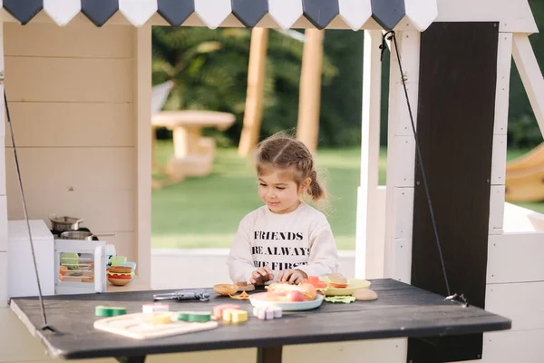Happy little girl playing on toy kitchen on wheels. Cute girl make a burger on toy kitchen outdoors. Adorable kid play on fresh air — Stock Fotó
