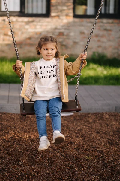 Happy little girl swing on a swing in the specially designated place in the park. Softner from sawdust underfoot — Stockfoto