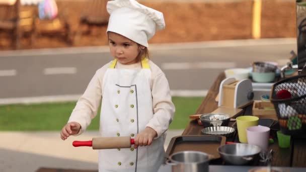 Adorable little girl in chefs coat and cap cooks at the childrens toy kitchen. Playing on little kids city. Start cooking — Stockvideo