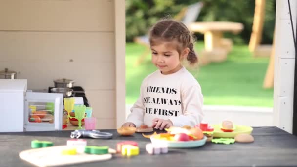 Happy little girl playing on toy kitchen on wheels. Cute girl make a burger on toy kitchen outdoors. Adorable kid play on fresh air — Wideo stockowe