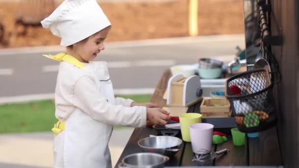 Adorable little girl in chefs coat and cap cooks at the childrens toy kitchen. Playing on little kids city. Start cooking — Stock Video