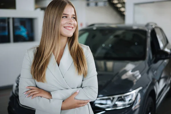 Portrait of salesperson in car showroom. Attractive young woman stend in front of car. Beautiful woman in suit in front of luxury car — Stock fotografie