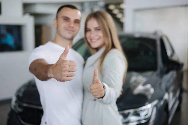 Casal jovem feliz depois de comprar carro novo do showroom do carro. Homem e mulher, levantem o polegar. Foco nas mãos — Fotografia de Stock