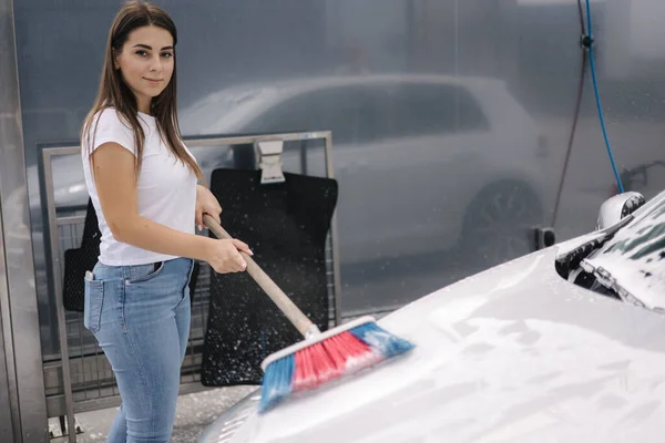 Attractive young woman washing her car with shampoo and brushes. Female washes automobile with foam and water outside on self service car wash.